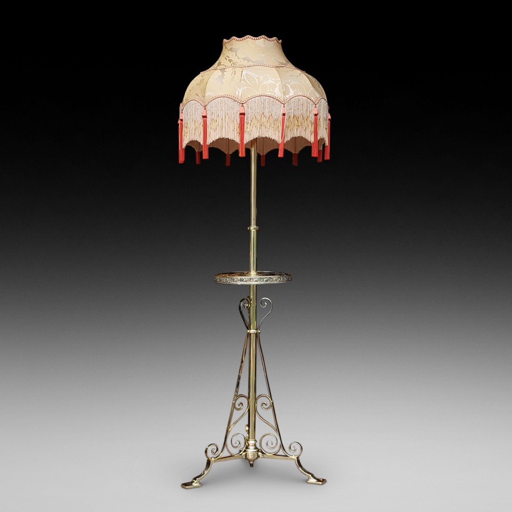 An Edwardian brass standard floor lamp featuring a scrollwork tripod base, an integrated circular tray table, and a fabric shade with fringe and tassels.