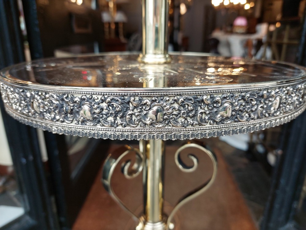 An Edwardian brass standard floor lamp featuring a scrollwork tripod base, an integrated circular tray table, and a fabric shade with fringe and tassels. View VI