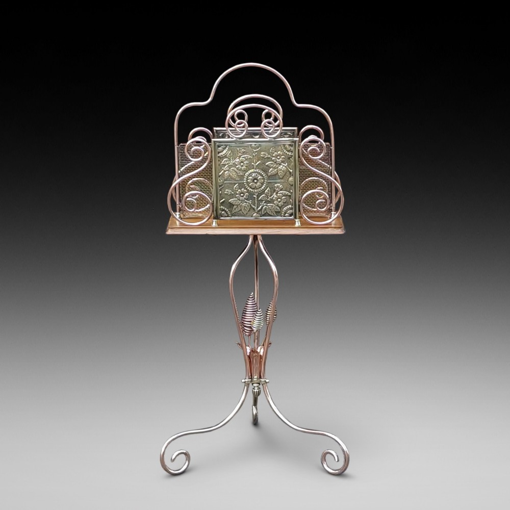 An Arts and Crafts copper and brass standing magazine rack featuring embossed floral metal panels, decorative wire scrollwork, and a tripod base with curved feet.