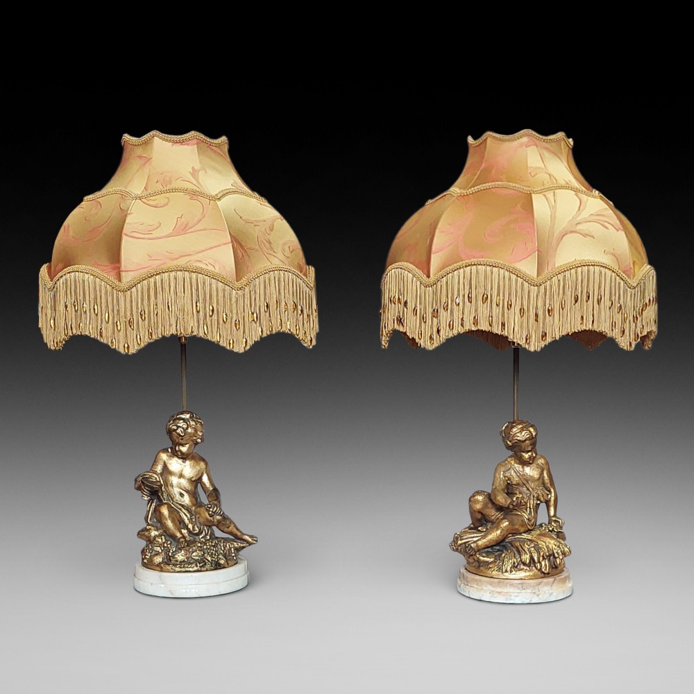 A pair of ornate table lamps featuring gilded cherub figures mounted on white alabaster bases, topped with fringed dome fabric shades.