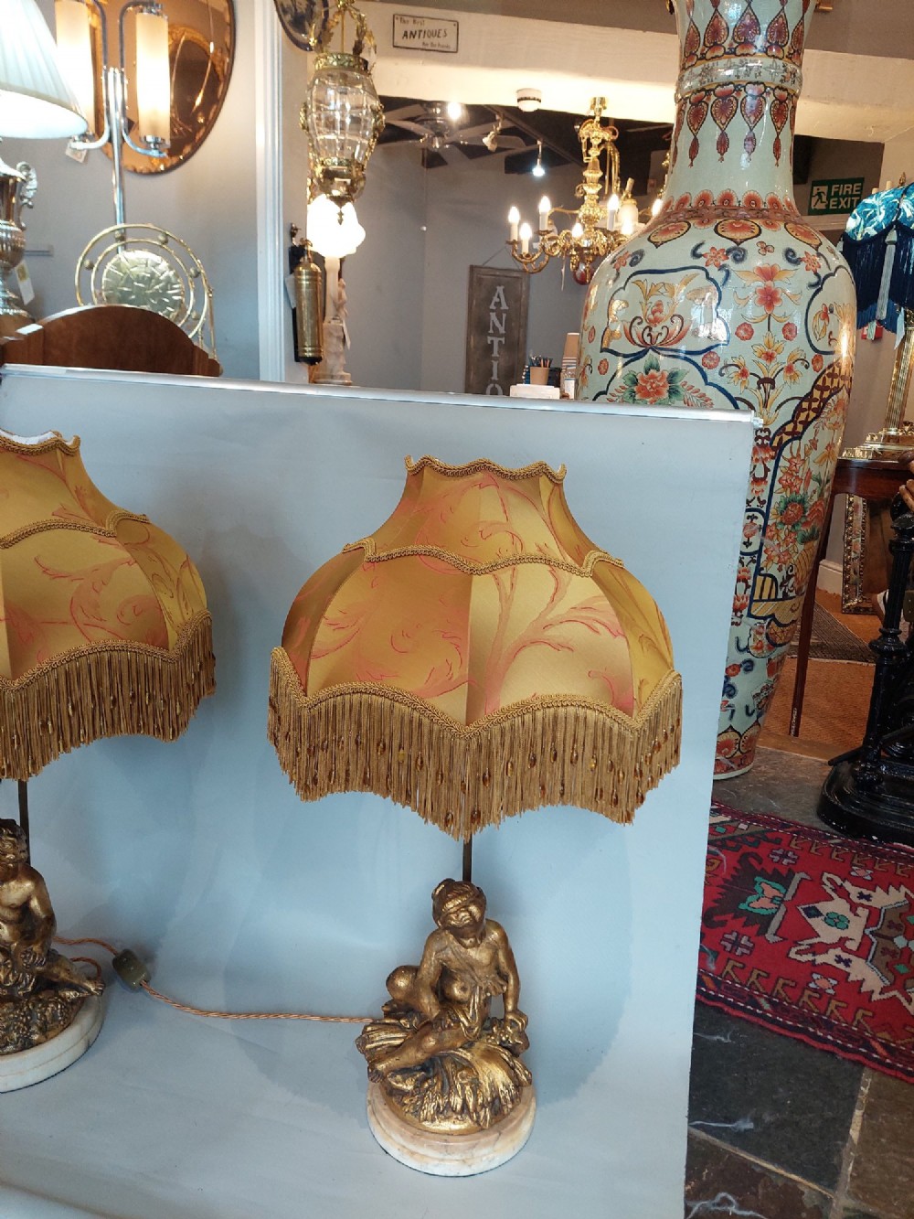 A pair of ornate table lamps featuring gilded cherub figures mounted on white alabaster bases, topped with fringed dome fabric shades. View II