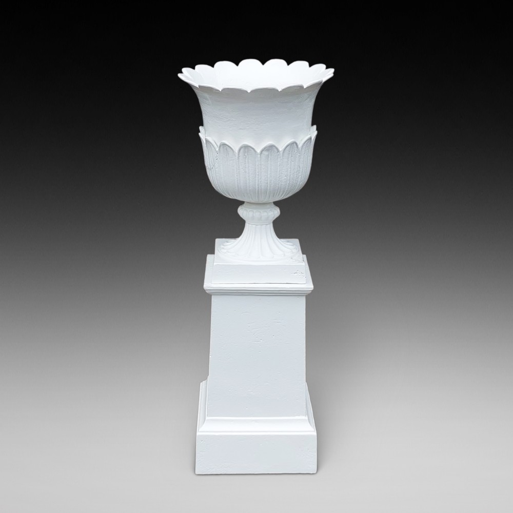 A classical white painted garden urn featuring an acanthus leaf pattern and scalloped rim, positioned on a matching square pedestal stand.