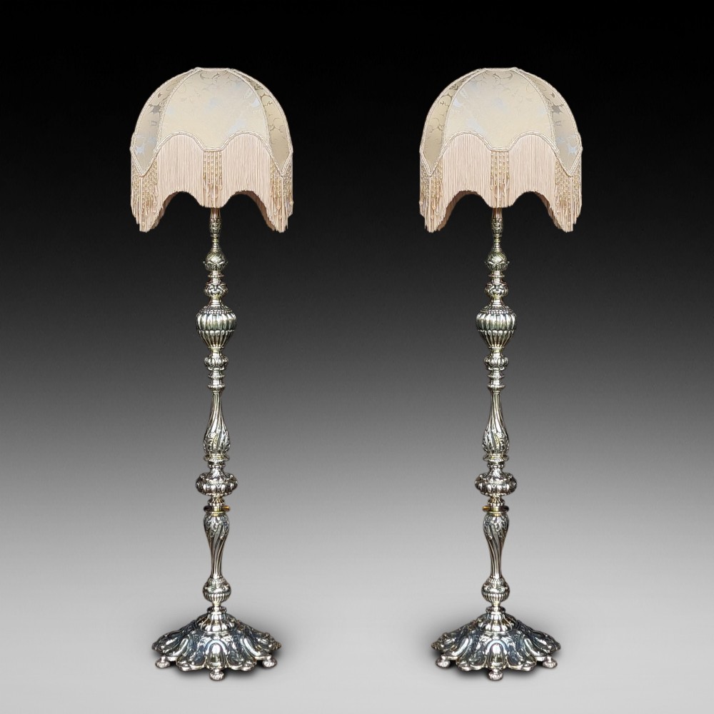 A pair of ornate silvered brass standard lamps featuring detailed turned baluster stems, lobed bases, and cream domed fabric shades with fringed edging.