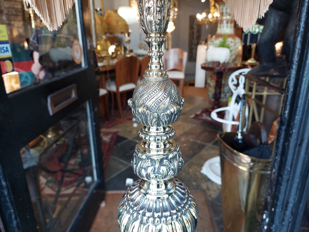 A pair of ornate silvered brass standard lamps featuring detailed turned baluster stems, lobed bases, and cream domed fabric shades with fringed edging. View X