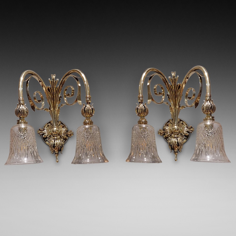 A pair of Edwardian cast brass double-arm wall sconces featuring leaf backplates and cut glass bell-shaped shades.
