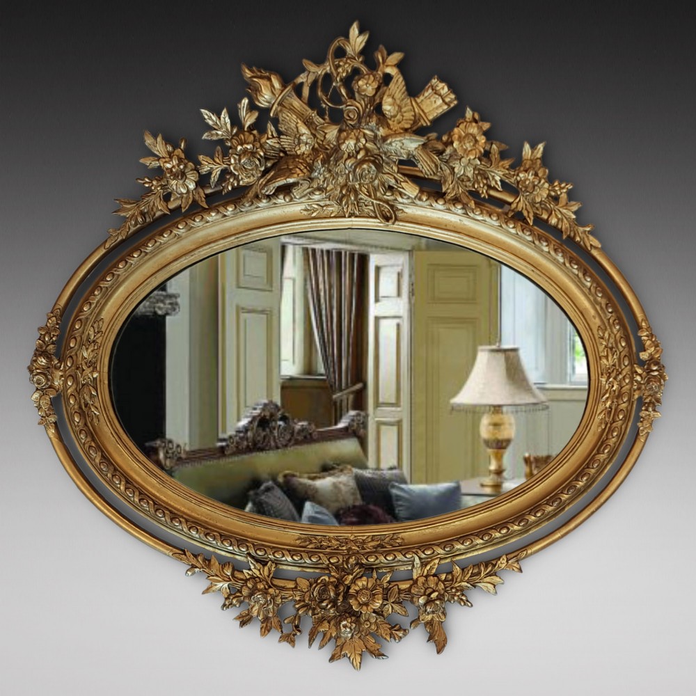 An oval 19th-century carved giltwood wall mirror featuring an intricate top crest with a torch and floral motif and an egg-and-dart inner frame border.