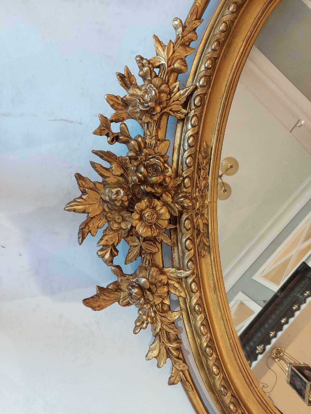 An oval 19th-century carved giltwood wall mirror featuring an intricate top crest with a torch and floral motif and an egg-and-dart inner frame border. View II