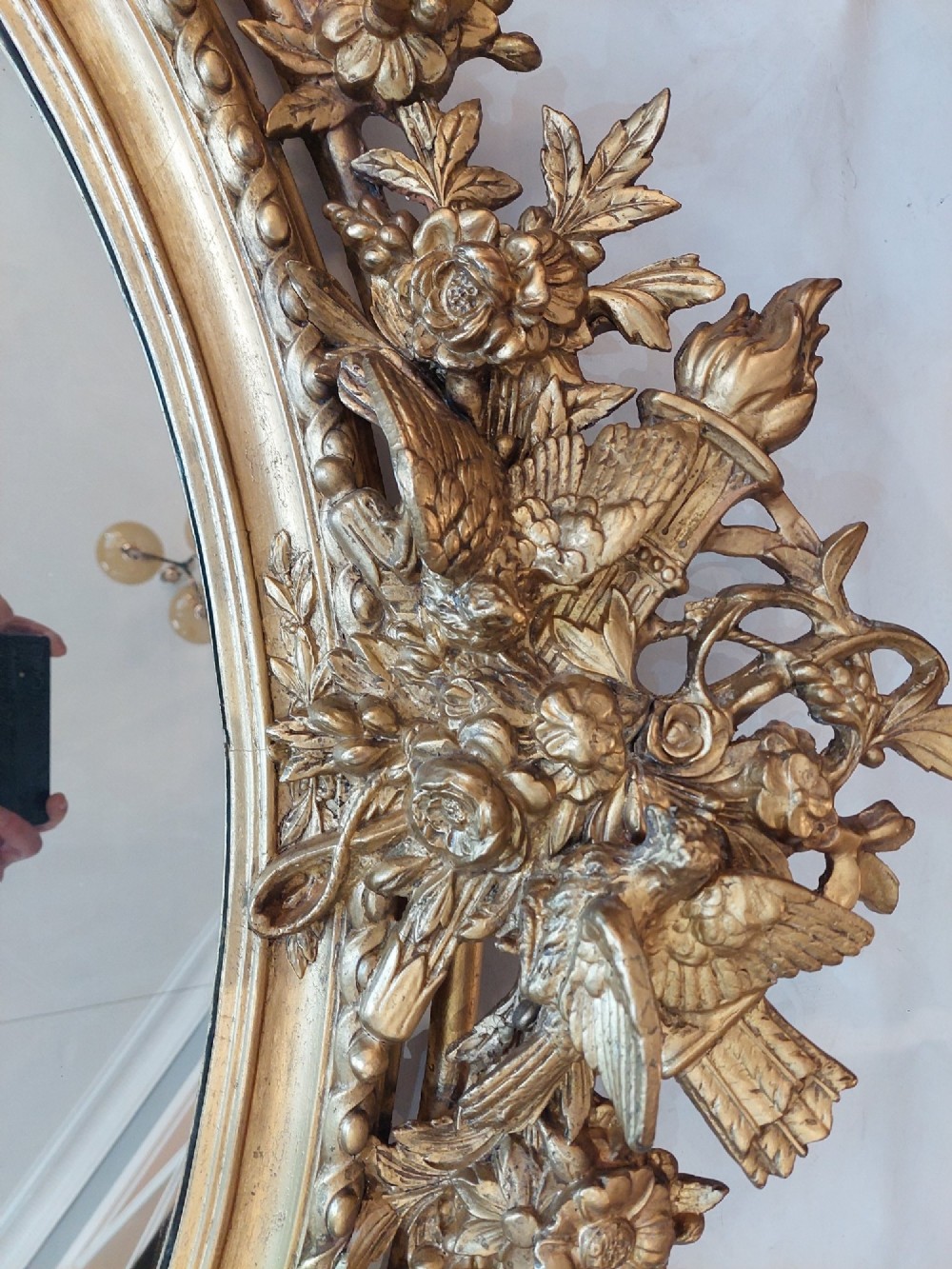An oval 19th-century carved giltwood wall mirror featuring an intricate top crest with a torch and floral motif and an egg-and-dart inner frame border. View IV