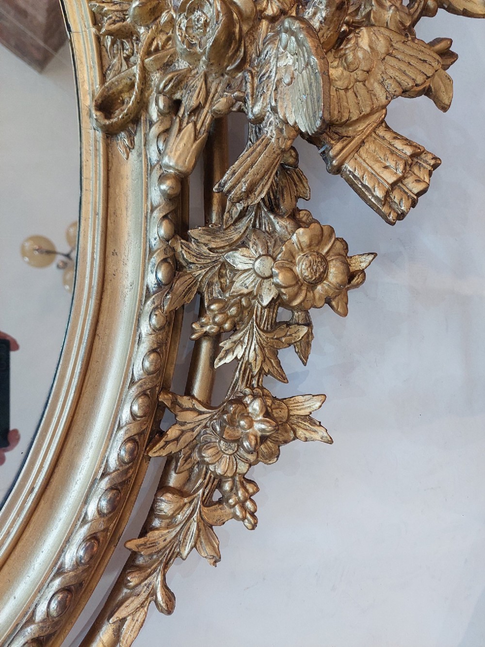 An oval 19th-century carved giltwood wall mirror featuring an intricate top crest with a torch and floral motif and an egg-and-dart inner frame border. View V