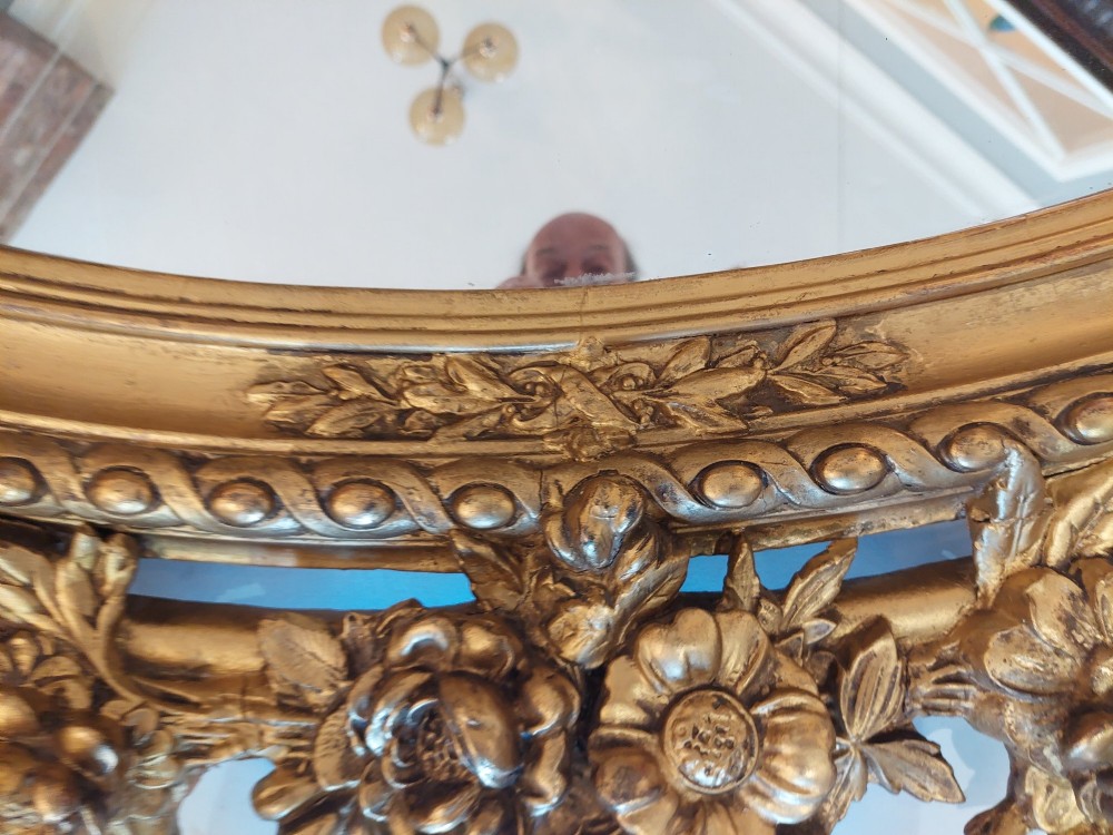 An oval 19th-century carved giltwood wall mirror featuring an intricate top crest with a torch and floral motif and an egg-and-dart inner frame border. View VI