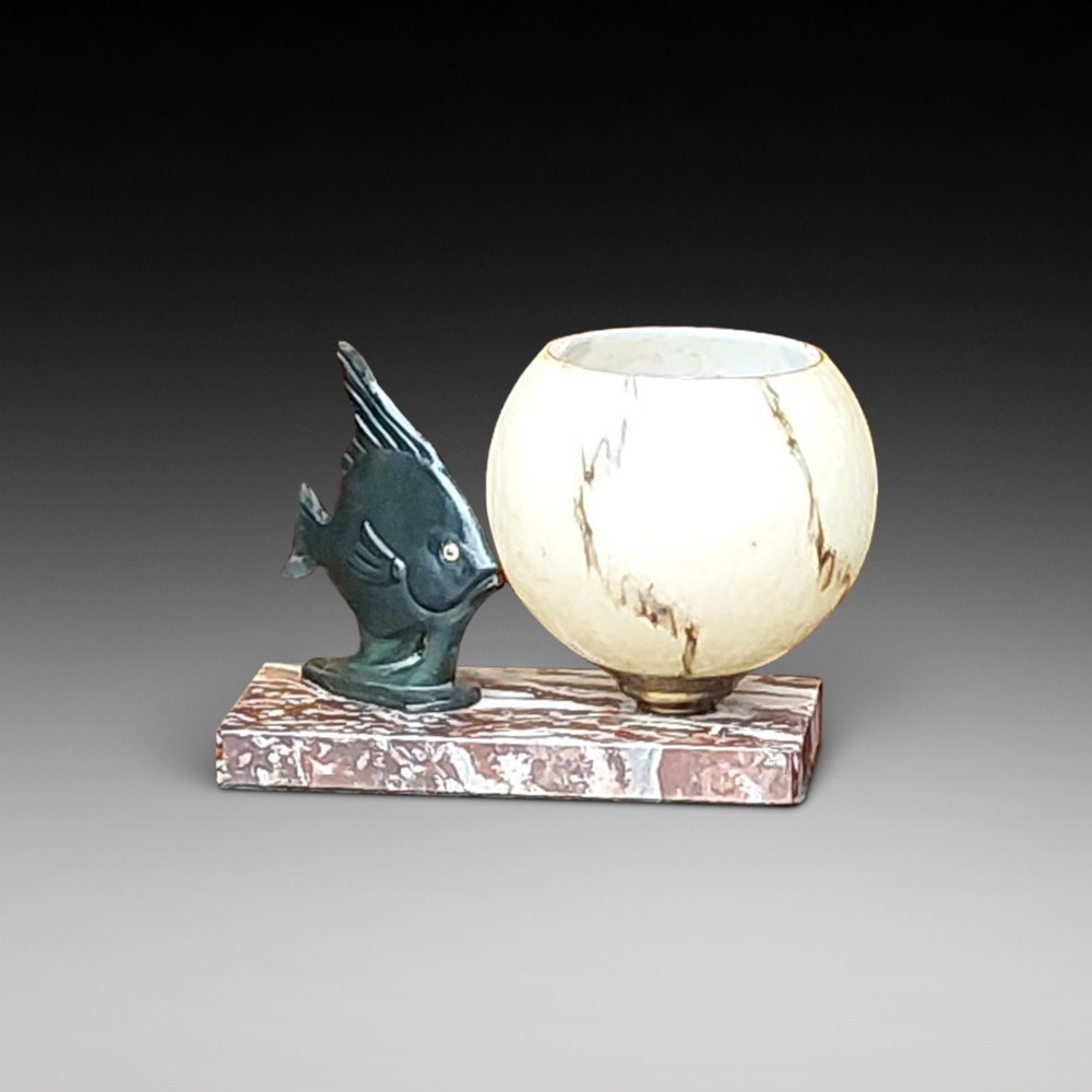 An Art Deco table lamp featuring a dark green patinated fish sculpture alongside a spherical cream glass shade, set on a rectangular variegated marble base.