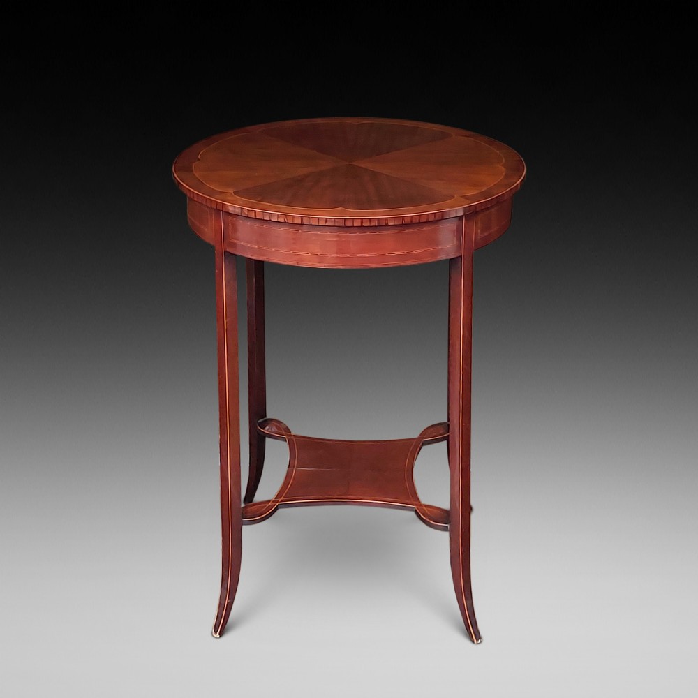 A circular Edwardian mahogany occasional table featuring a segmented veneer top with boxwood stringing inlay, splayed legs, and a lower concave stretcher shelf.