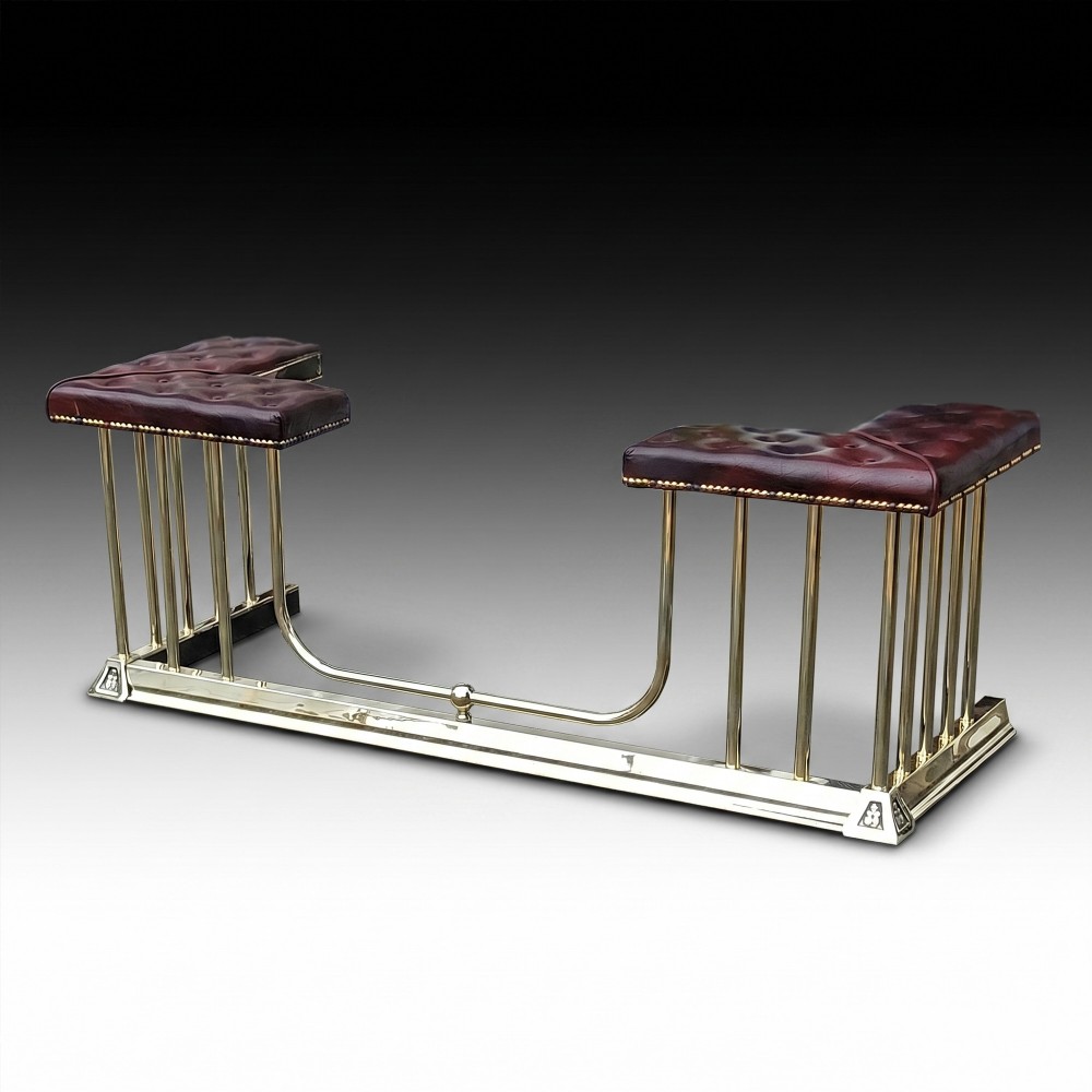 An early 20th-century brass club fender featuring a central dipped rail, vertical brass balusters, and two button-tufted dark red leather upholstered corner seats.