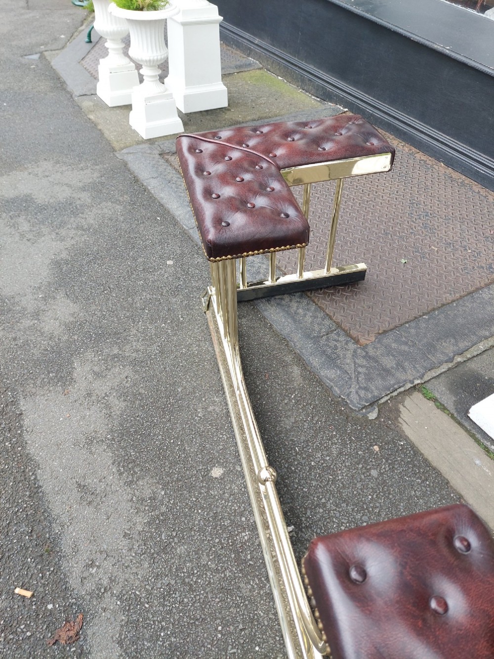 An early 20th-century brass club fender featuring a central dipped rail, vertical brass balusters, and two button-tufted dark red leather upholstered corner seats. View IV