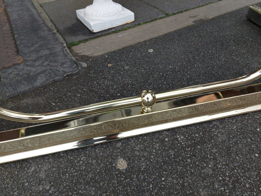 An early 20th-century brass club fender featuring a central dipped rail, vertical brass balusters, and two button-tufted dark red leather upholstered corner seats. View X