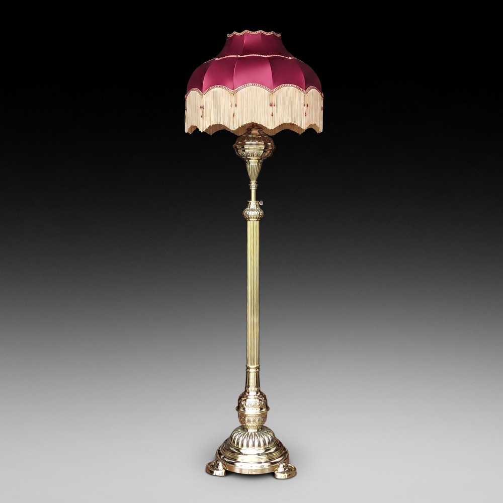 A Victorian brass extending standard floor lamp featuring a fluted column base and a deep red fabric shade with fringe detailing.