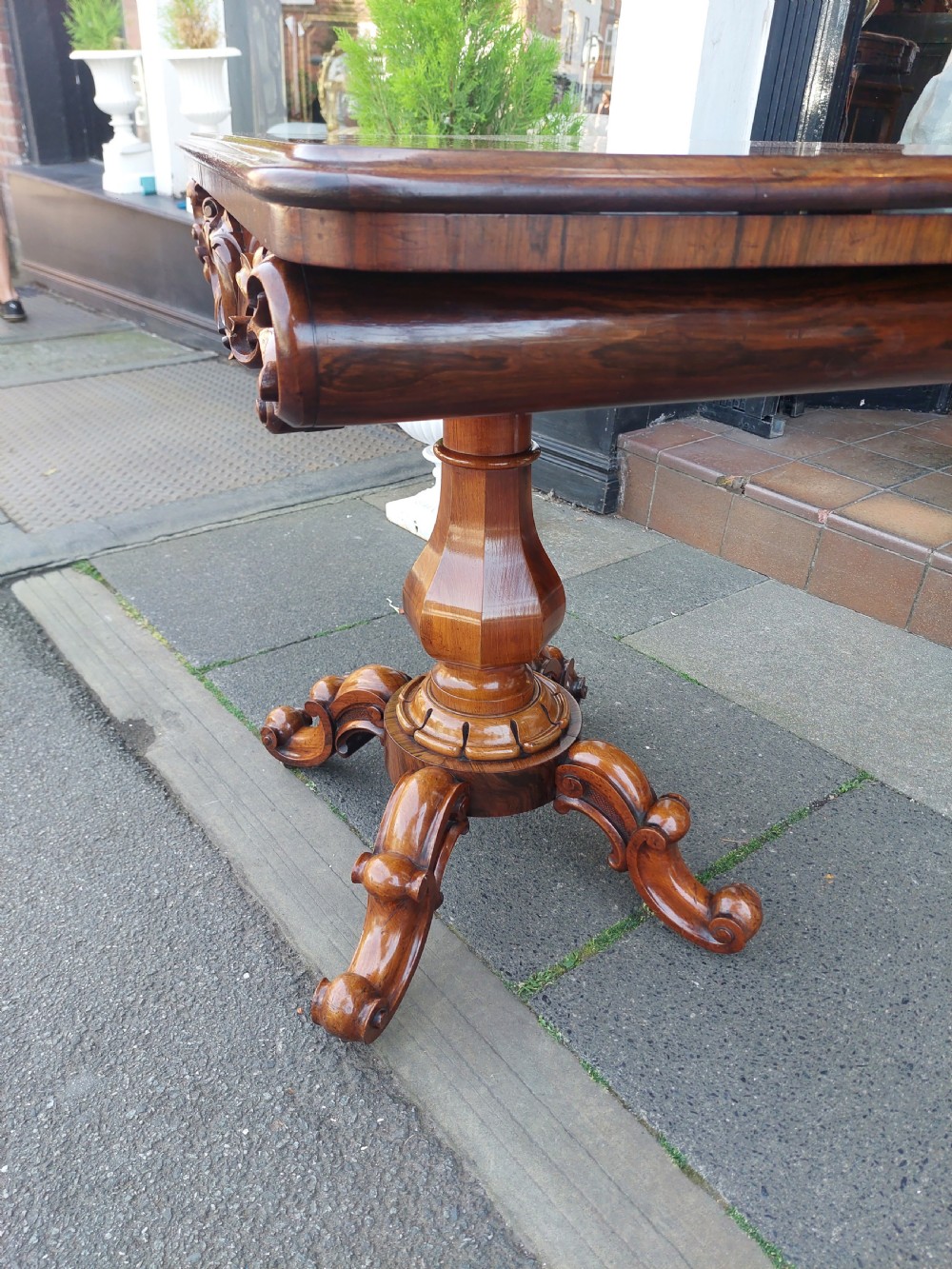 A 19th-century William IV rosewood tea table featuring a polished rectangular fold-over top, carved scrollwork frieze, bulbous faceted pedestal stem, and scroll feet on brass castors. View II