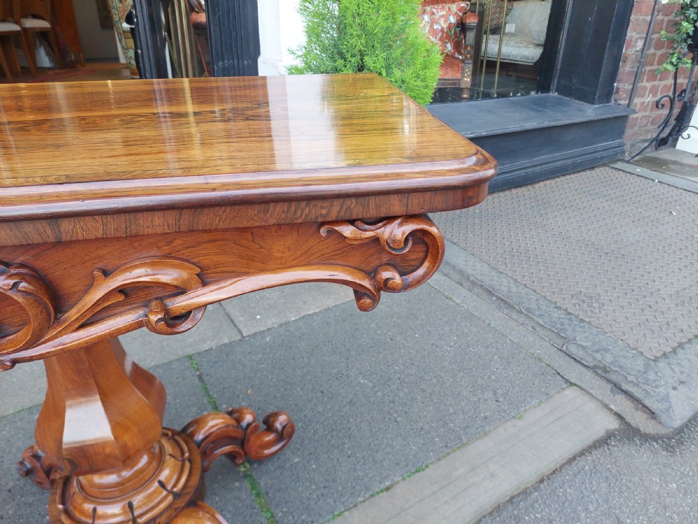 A 19th-century William IV rosewood tea table featuring a polished rectangular fold-over top, carved scrollwork frieze, bulbous faceted pedestal stem, and scroll feet on brass castors. View V