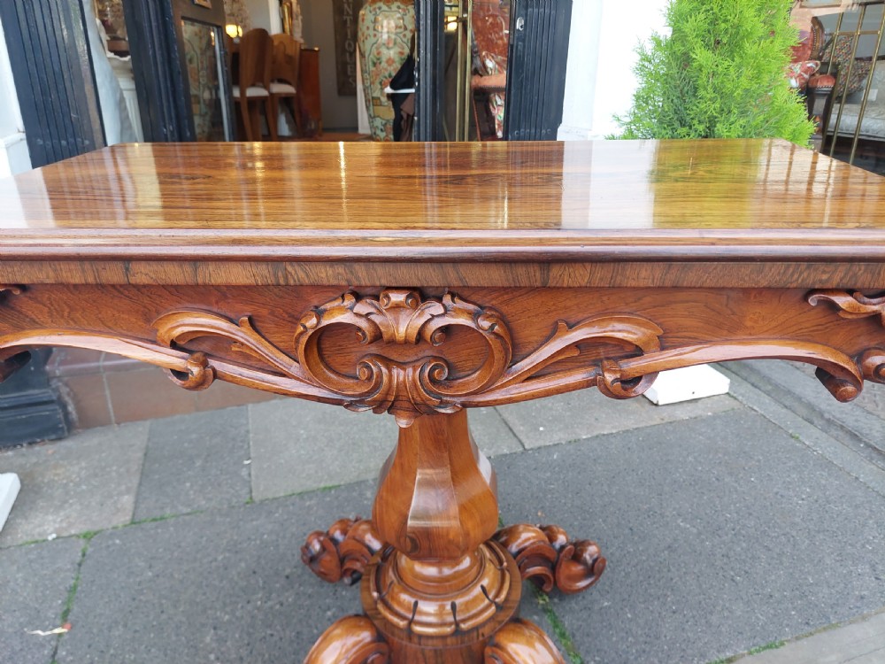 A 19th-century William IV rosewood tea table featuring a polished rectangular fold-over top, carved scrollwork frieze, bulbous faceted pedestal stem, and scroll feet on brass castors. View VI