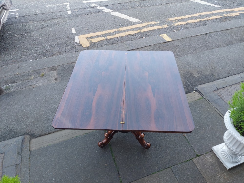 A 19th-century William IV rosewood tea table featuring a polished rectangular fold-over top, carved scrollwork frieze, bulbous faceted pedestal stem, and scroll feet on brass castors. View VII