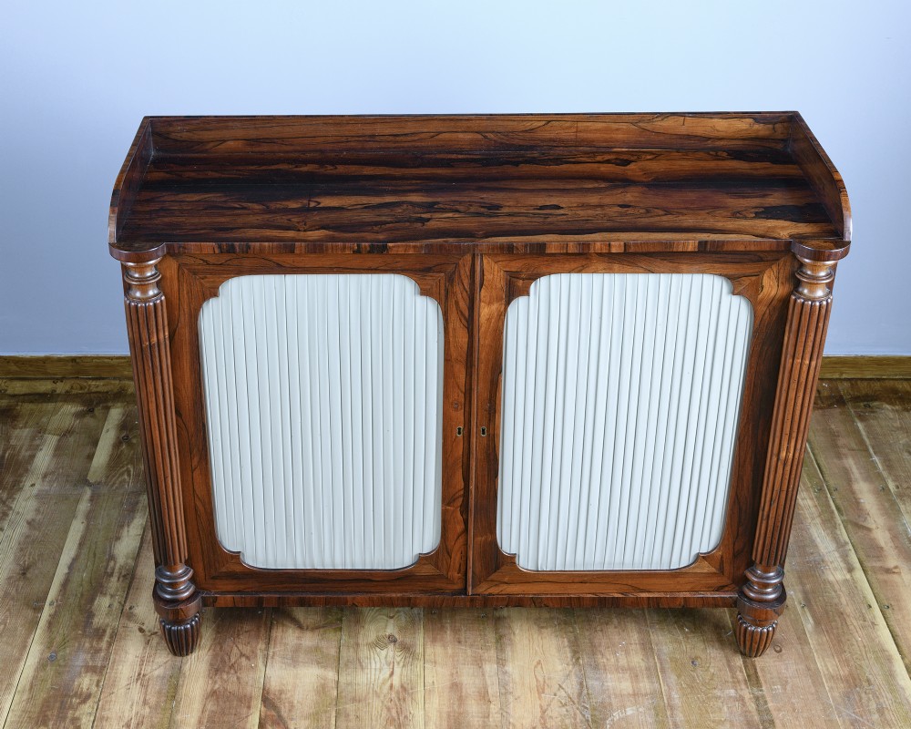 A Regency Goncalo Alves side cabinet featuring two pleated light fabric panel doors flanked by fluted columns on turned feet. View IV