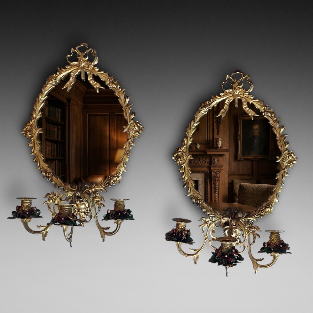 A pair of early 20th-century oval gilt metal wall mirrors with ribbon-tied crests and three candle branches.