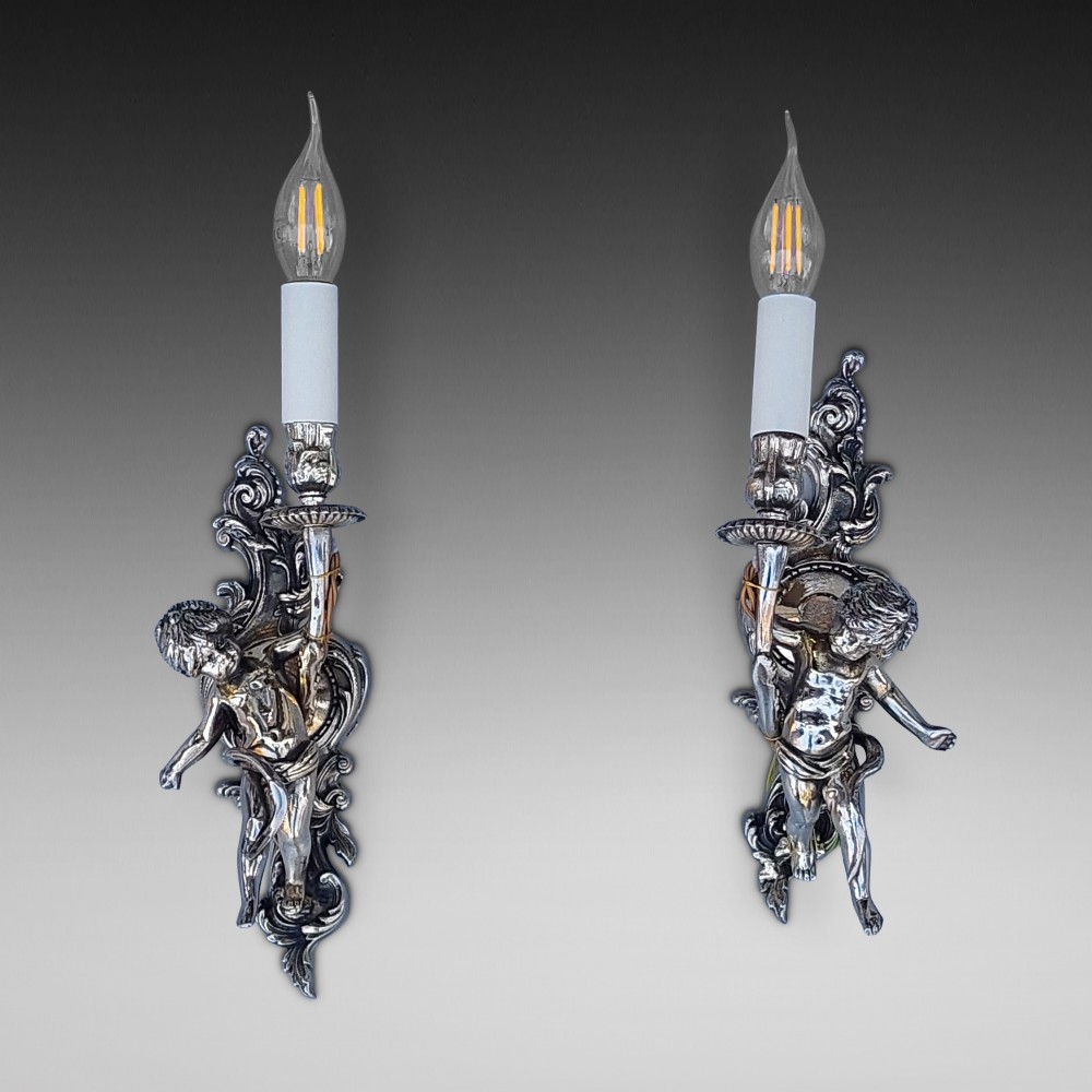 A pair of early 20th-century silvered metal wall sconces featuring sculpted cherubs holding single candle sockets set against elaborate scrollwork backplates.