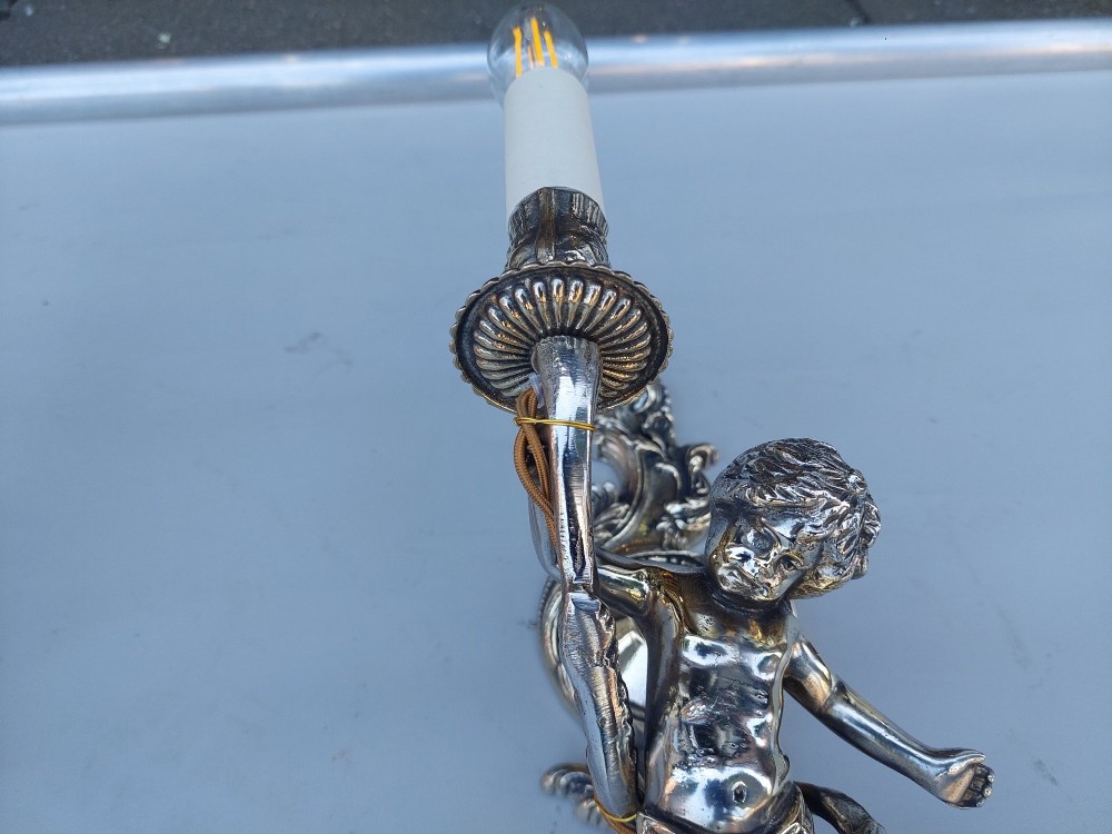 A pair of early 20th-century silvered metal wall sconces featuring sculpted cherubs holding single candle sockets set against elaborate scrollwork backplates. View III
