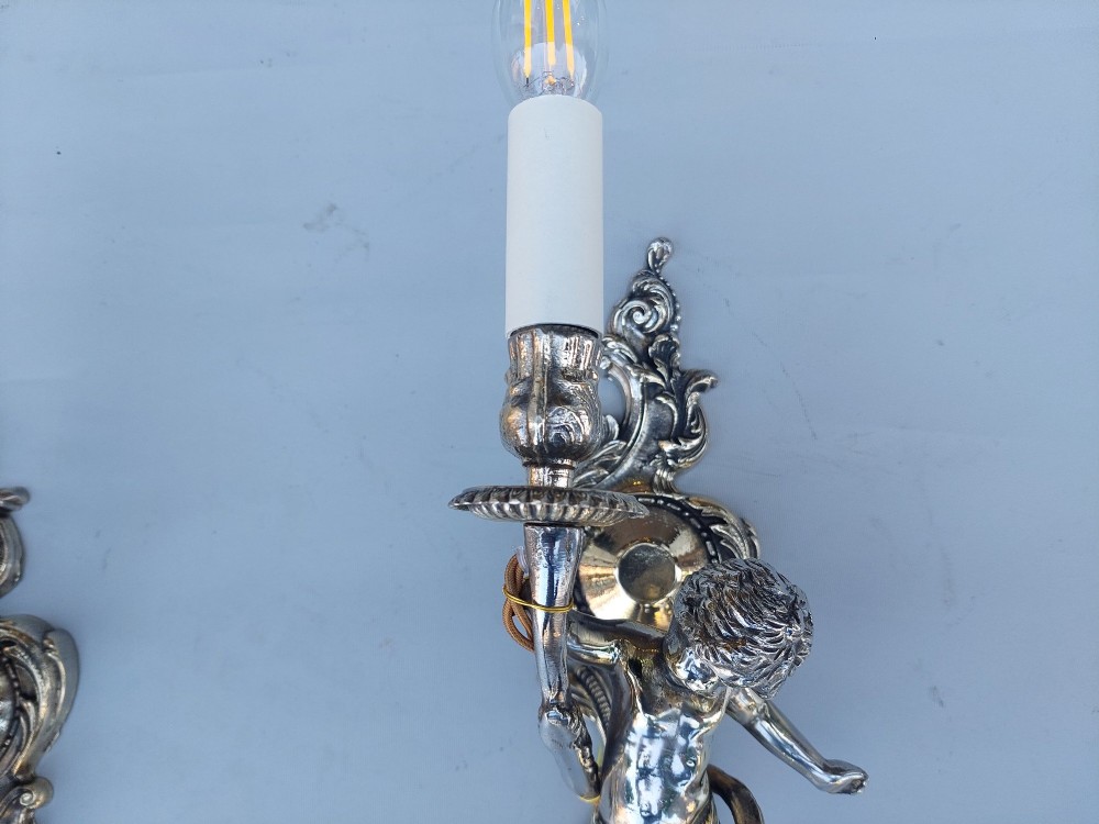 A pair of early 20th-century silvered metal wall sconces featuring sculpted cherubs holding single candle sockets set against elaborate scrollwork backplates. View IV