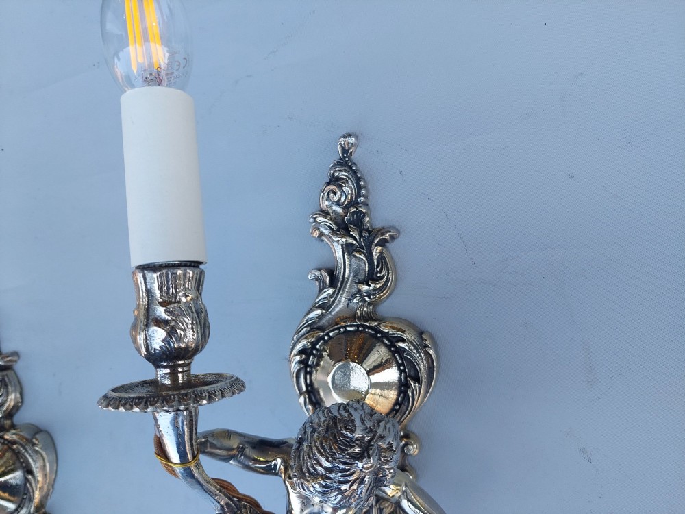 A pair of early 20th-century silvered metal wall sconces featuring sculpted cherubs holding single candle sockets set against elaborate scrollwork backplates. View V