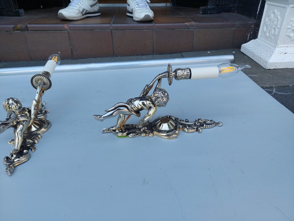 A pair of early 20th-century silvered metal wall sconces featuring sculpted cherubs holding single candle sockets set against elaborate scrollwork backplates. View VIII