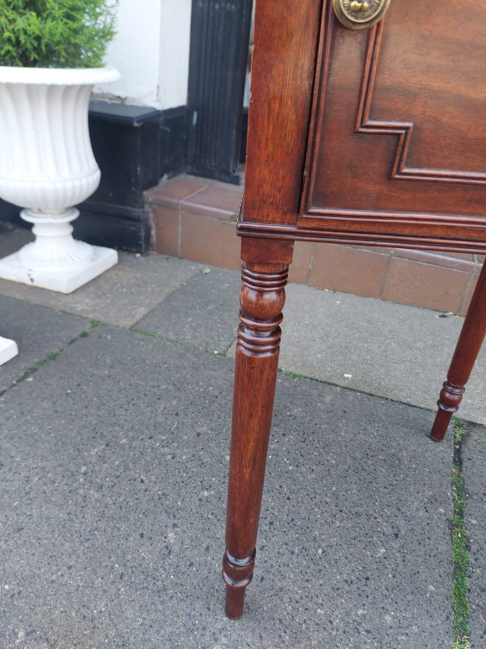 An early 19th-century Regency mahogany bedside cabinet featuring a single panelled door, tray top gallery, turned legs, and a brass knob. View V