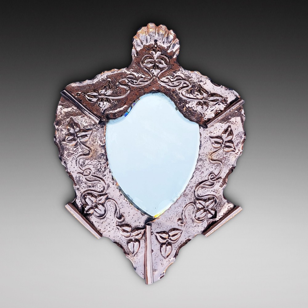 A shield-shaped wall mirror featuring a hand-hammered copper frame decorated with floral repousse motifs and a bevelled glass centre.