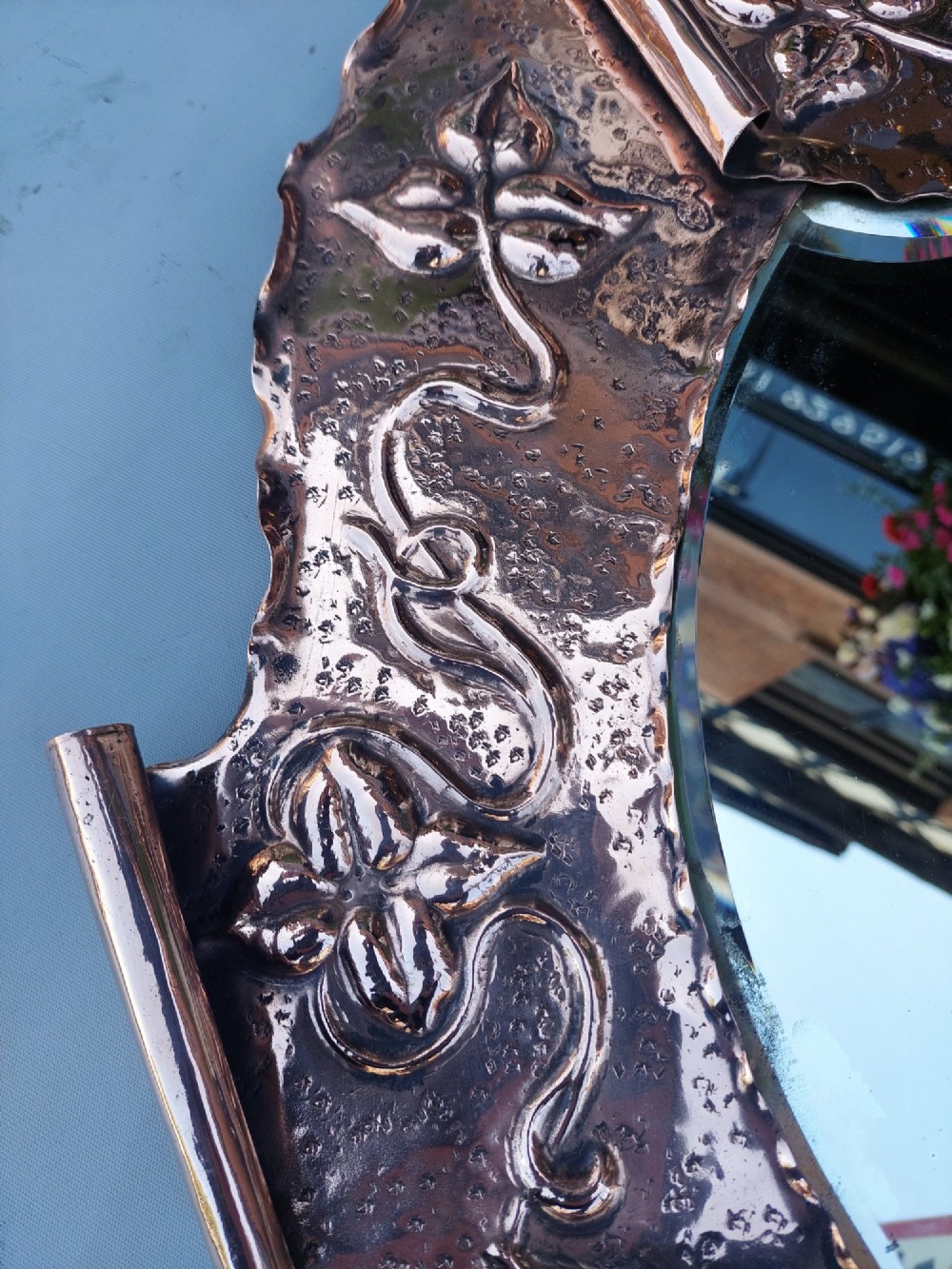 A shield-shaped wall mirror featuring a hand-hammered copper frame decorated with floral repousse motifs and a bevelled glass centre. View III