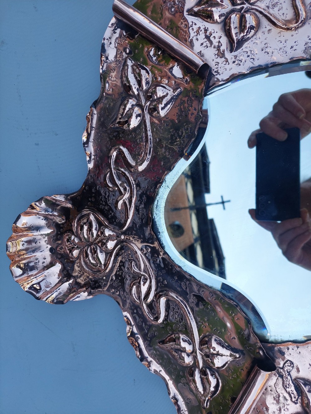 A shield-shaped wall mirror featuring a hand-hammered copper frame decorated with floral repousse motifs and a bevelled glass centre. View V