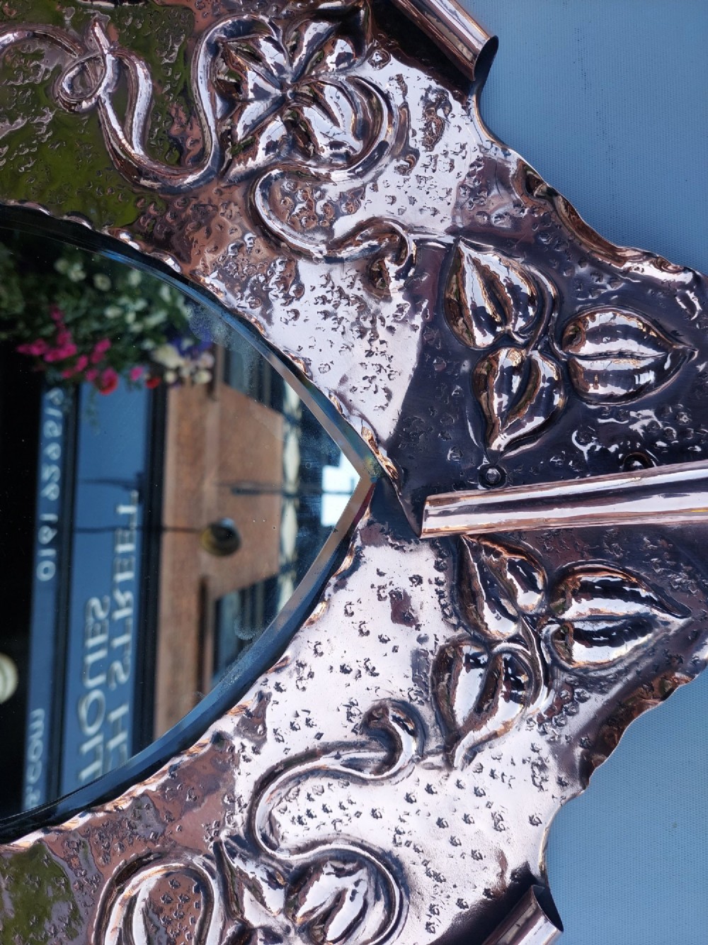 A shield-shaped wall mirror featuring a hand-hammered copper frame decorated with floral repousse motifs and a bevelled glass centre. View VI