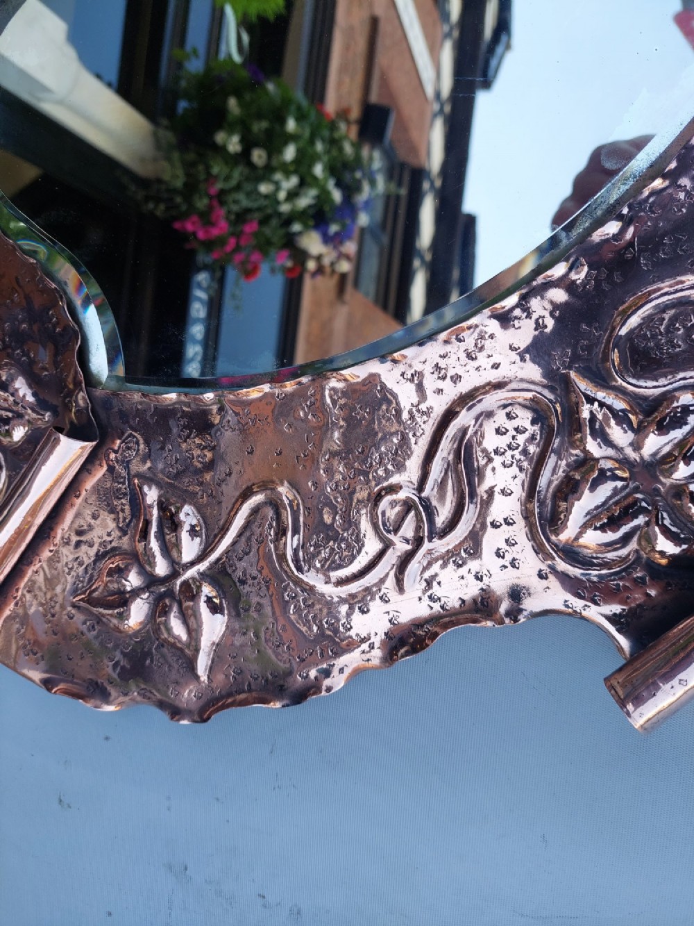 A shield-shaped wall mirror featuring a hand-hammered copper frame decorated with floral repousse motifs and a bevelled glass centre. View VII