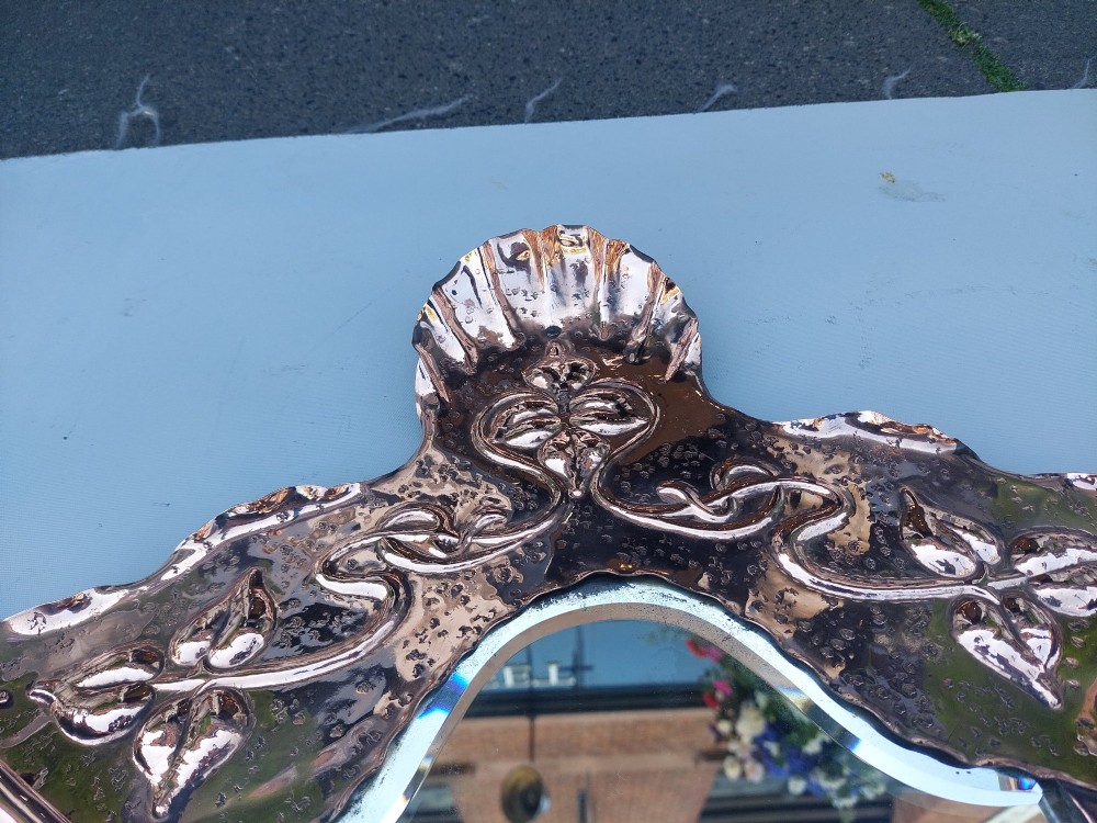 A shield-shaped wall mirror featuring a hand-hammered copper frame decorated with floral repousse motifs and a bevelled glass centre. View VIII
