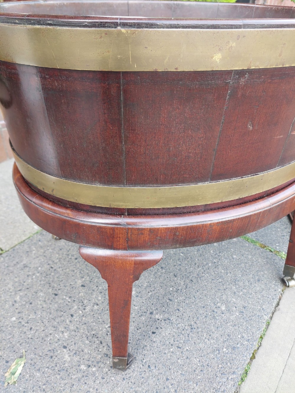 An 18th-century oval dark mahogany wine cooler with brass binding straps and carrying handles, elevated on a four-legged stand with castors. View IX