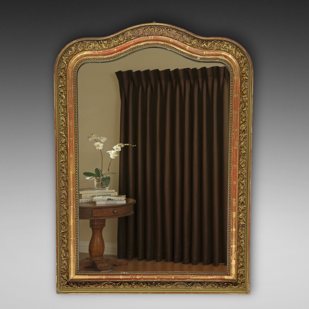 A late nineteenth-century wall mirror featuring an arched giltwood frame with carved foliate details, a beaded inner border, and distressed gold leaf revealing underlying red bole.