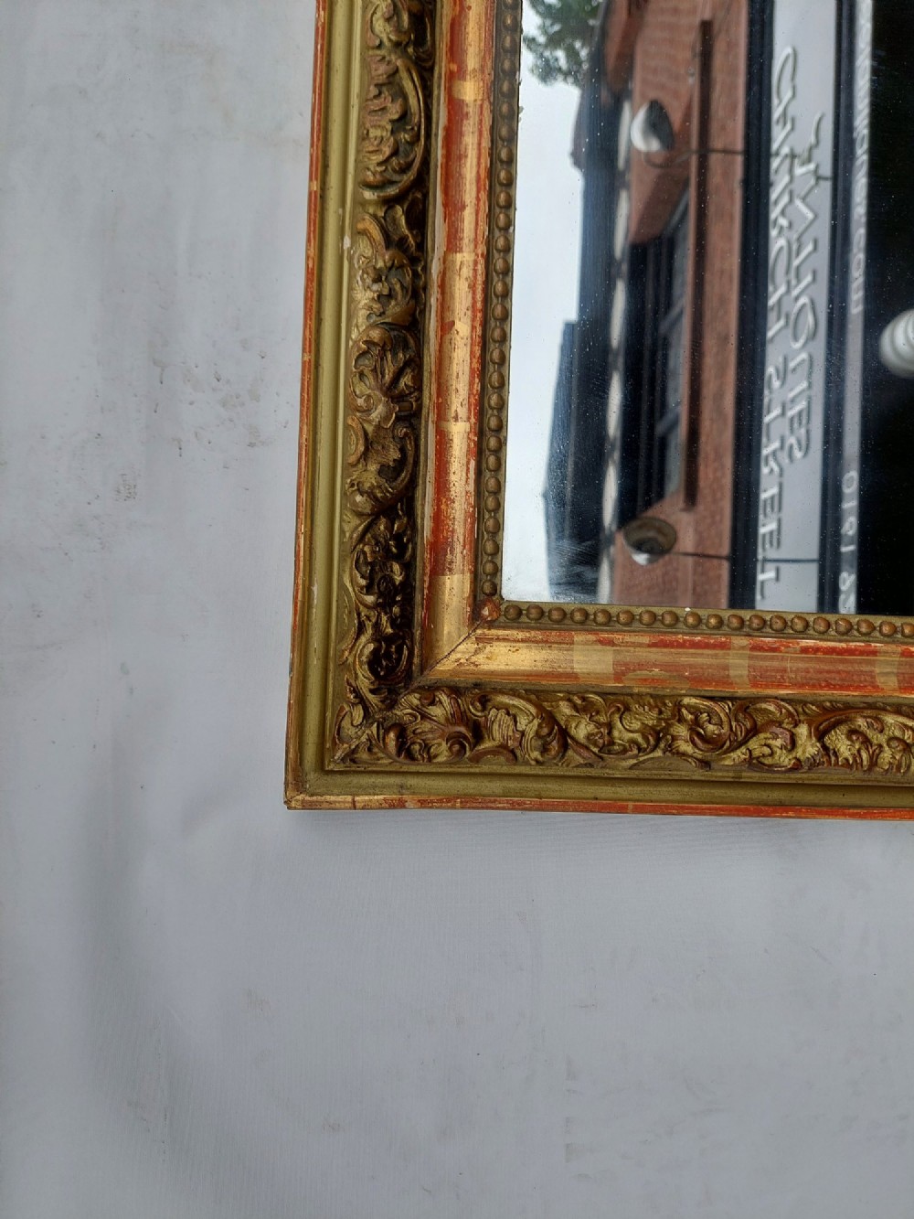A late nineteenth-century wall mirror featuring an arched giltwood frame with carved foliate details, a beaded inner border, and distressed gold leaf revealing underlying red bole. View II