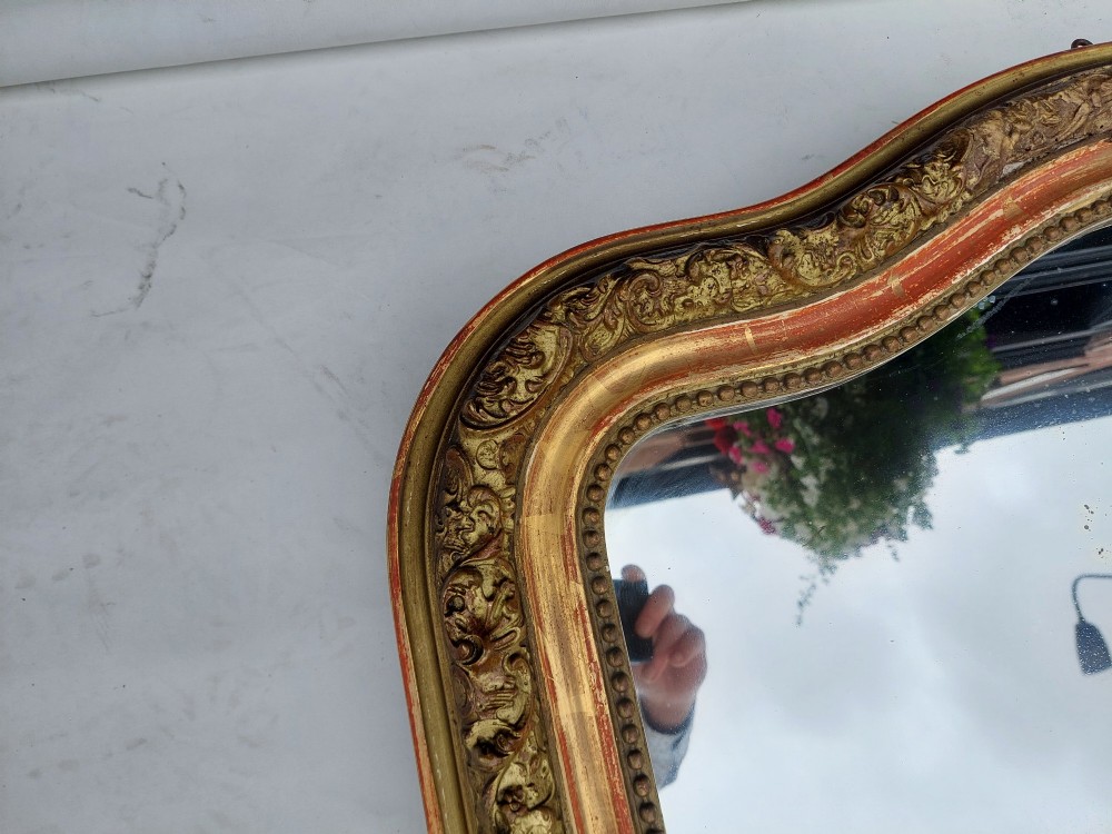 A late nineteenth-century wall mirror featuring an arched giltwood frame with carved foliate details, a beaded inner border, and distressed gold leaf revealing underlying red bole. View V