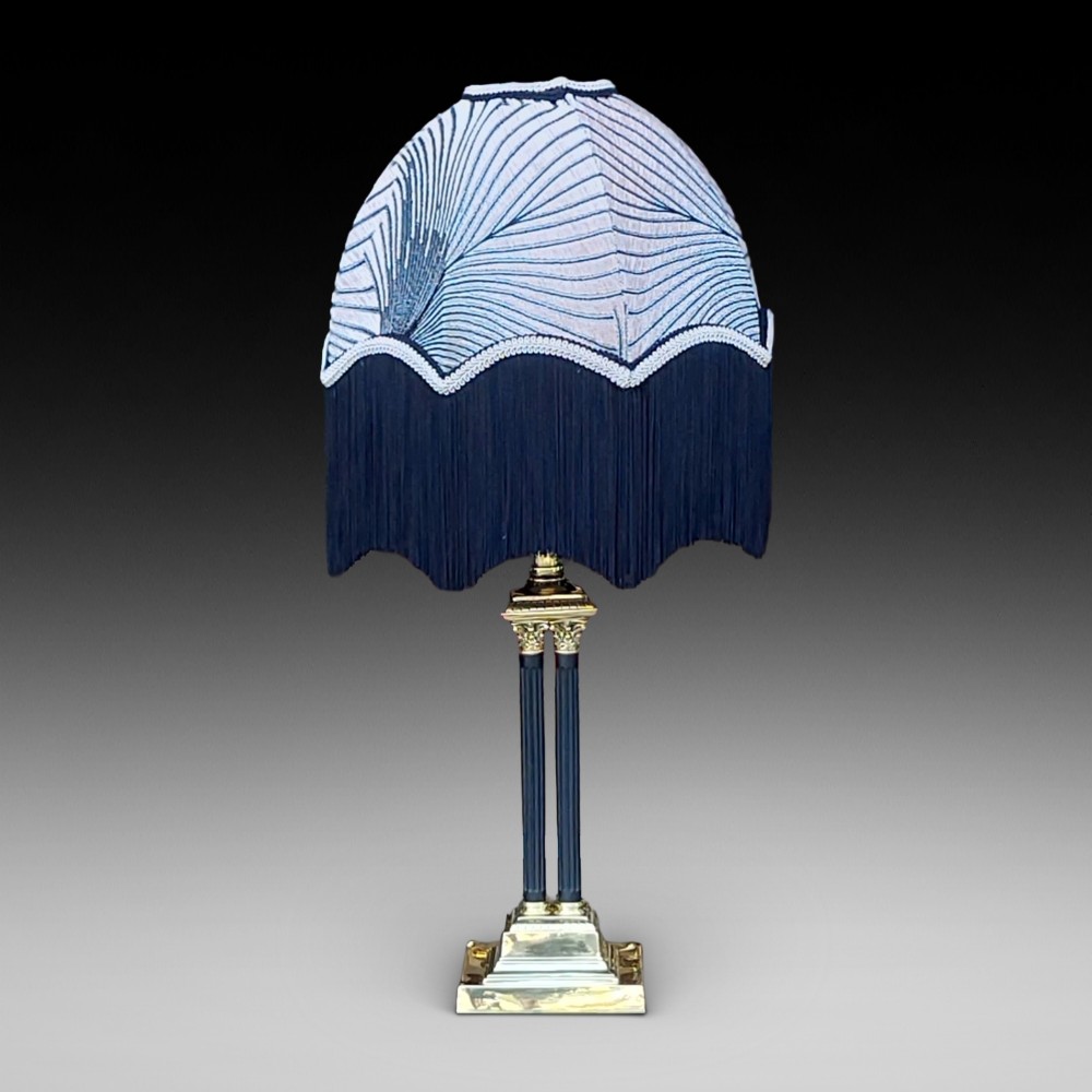 An antique brass table lamp featuring a four-pillar Corinthian column base and a patterned blue dome lampshade with dark fringe.