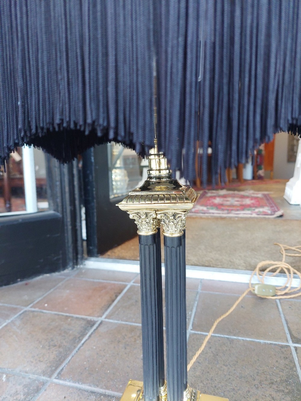 An antique brass table lamp featuring a four-pillar Corinthian column base and a patterned blue dome lampshade with dark fringe. View IV