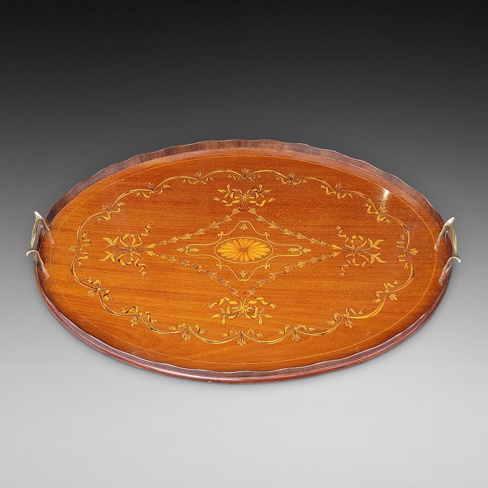 An oval mahogany tray featuring detailed floral and bow marquetry inlays, a scalloped gallery edge, and two brass side handles.