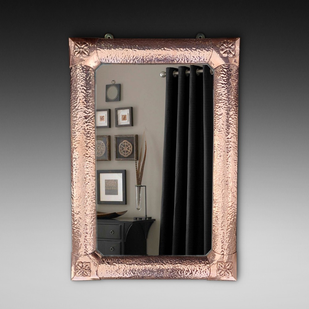 A rectangular wall mirror with a hand-beaten copper frame featuring raised floral rosettes at each corner.