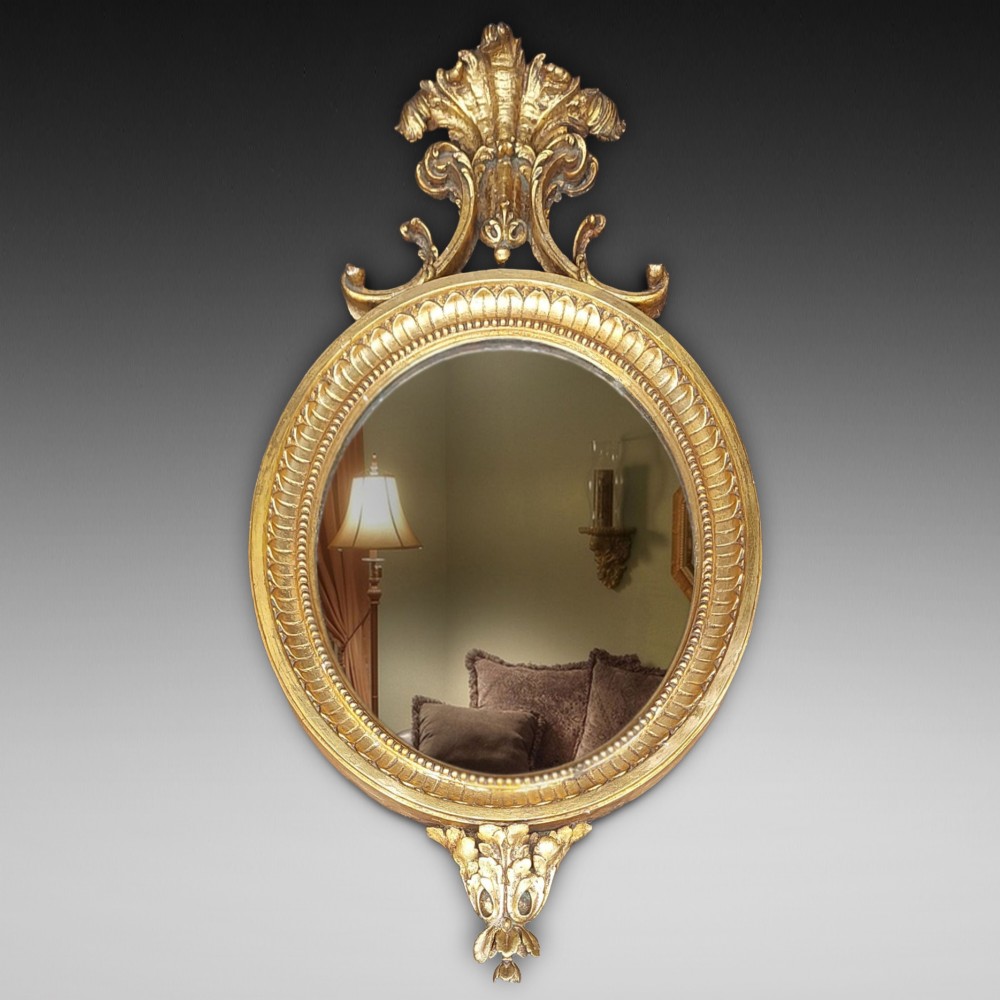 An early 19th-century oval giltwood wall mirror featuring a carved acanthus cresting and matching bottom pendant.