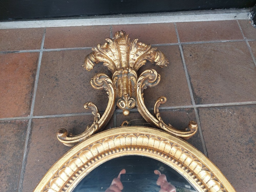 An early 19th-century oval giltwood wall mirror featuring a carved acanthus cresting and matching bottom pendant. View V