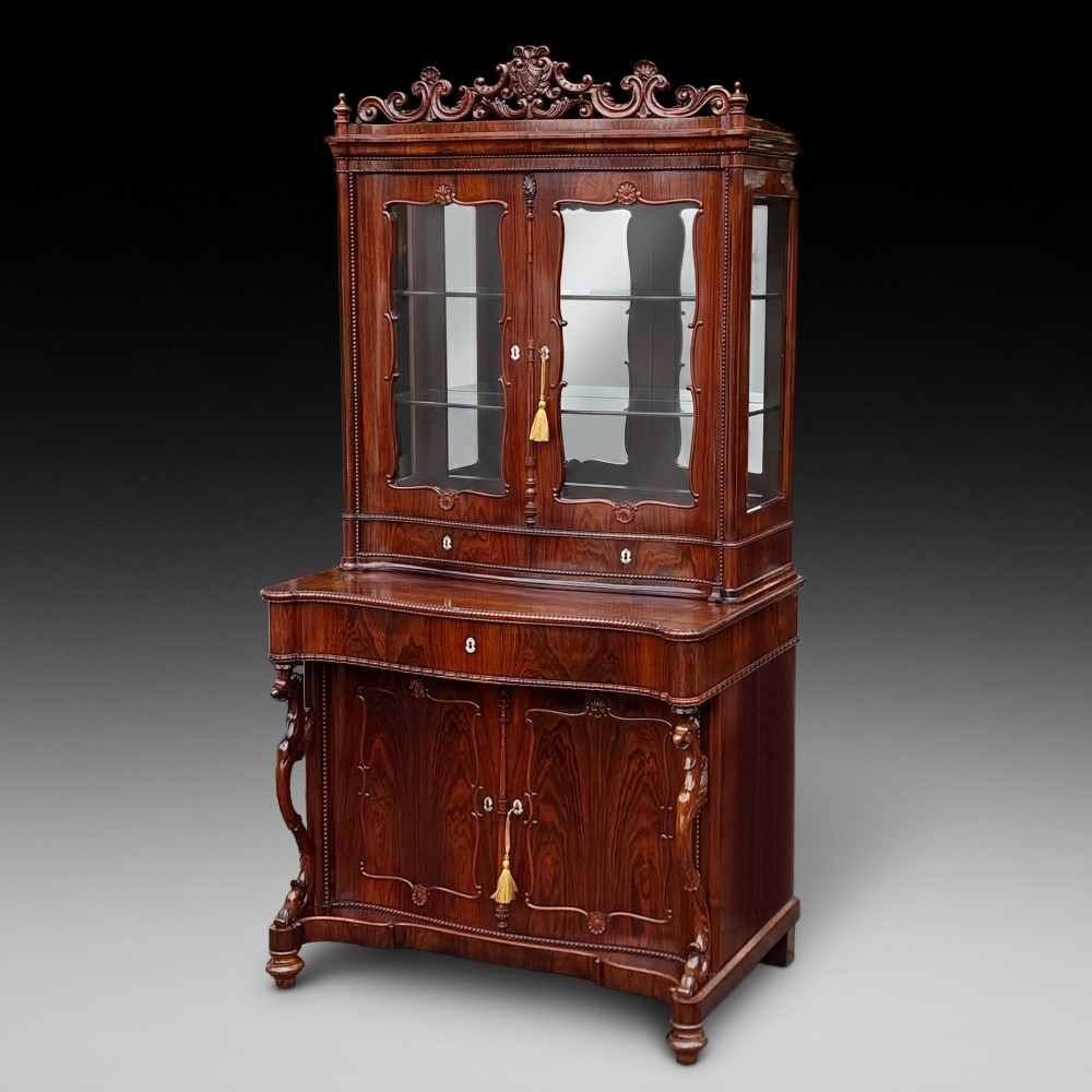 A two-tier Biedermeier rosewood display cabinet featuring a glazed upper section with a carved cresting above a two-door cupboard base.