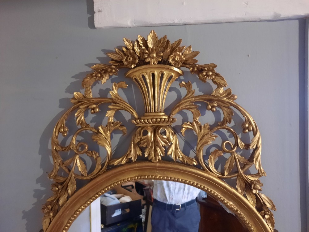 An oval giltwood wall mirror featuring an intricate carved urn cresting, acanthus leaf foliage, and a beaded inner frame border. View II